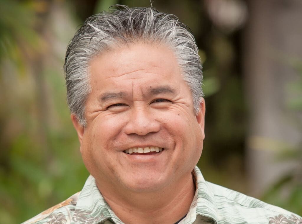 Smiling middle-aged man with short gray hair outdoors, wearing a tropical patterned shirt.