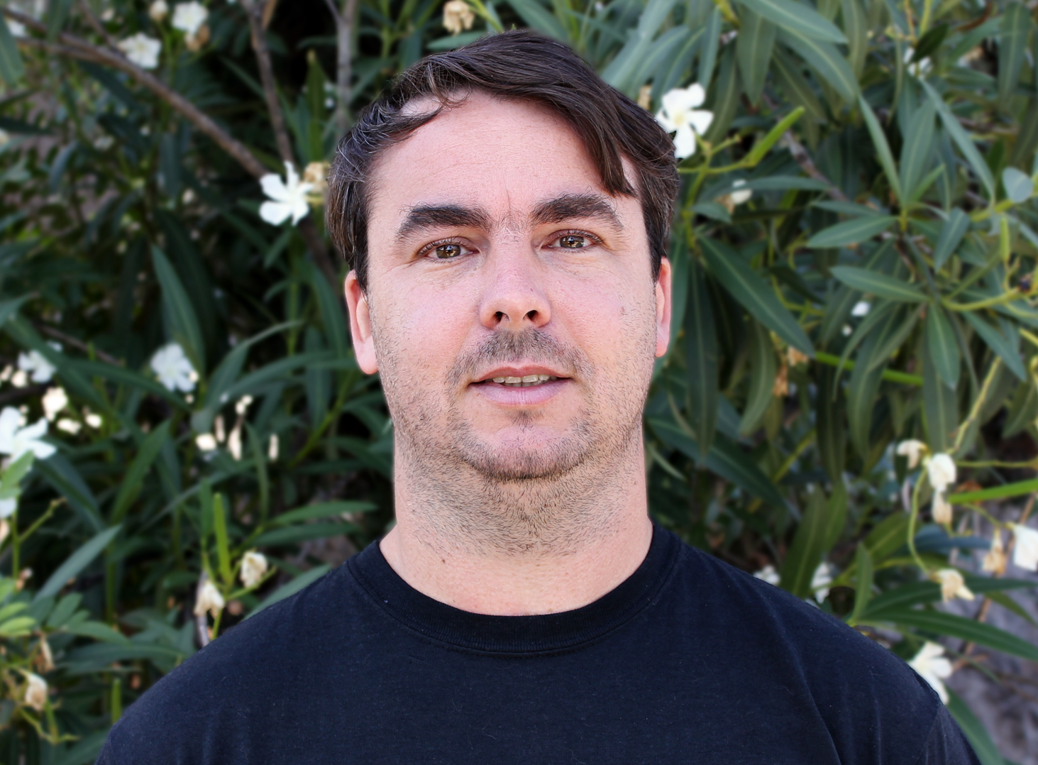 Portrait of a man with short dark hair wearing a black shirt, standing in front of green foliage with white flowers.
