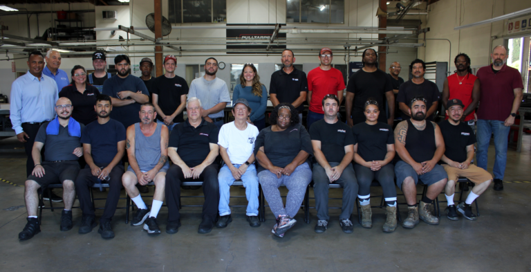 Group of factory workers posing for a team photo in a workshop, with equipment and overhead rails in the background.