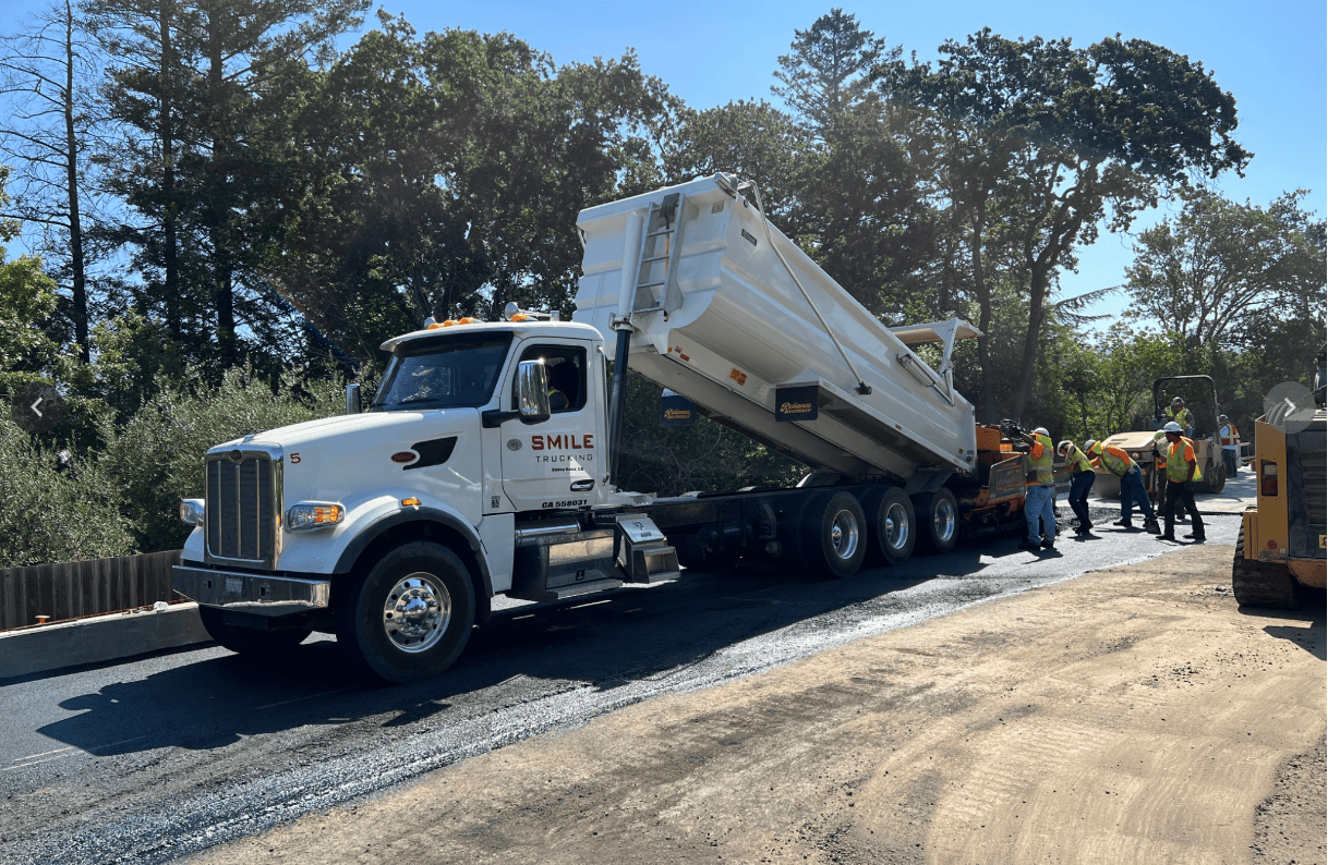 White dump truck labeled Smile Trucking on a road construction site, dump bed raised as material is unloaded, with workers in safety vests nearby.