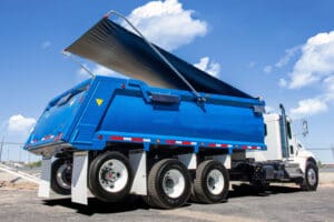 Blue dump truck with raised cargo bed on a construction site under a sunny sky with few clouds.